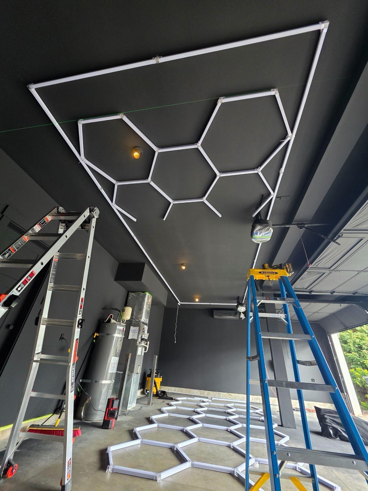Hexagon LED light frame being mounted on a dark garage ceiling, with more hex frames laid out on the slab below