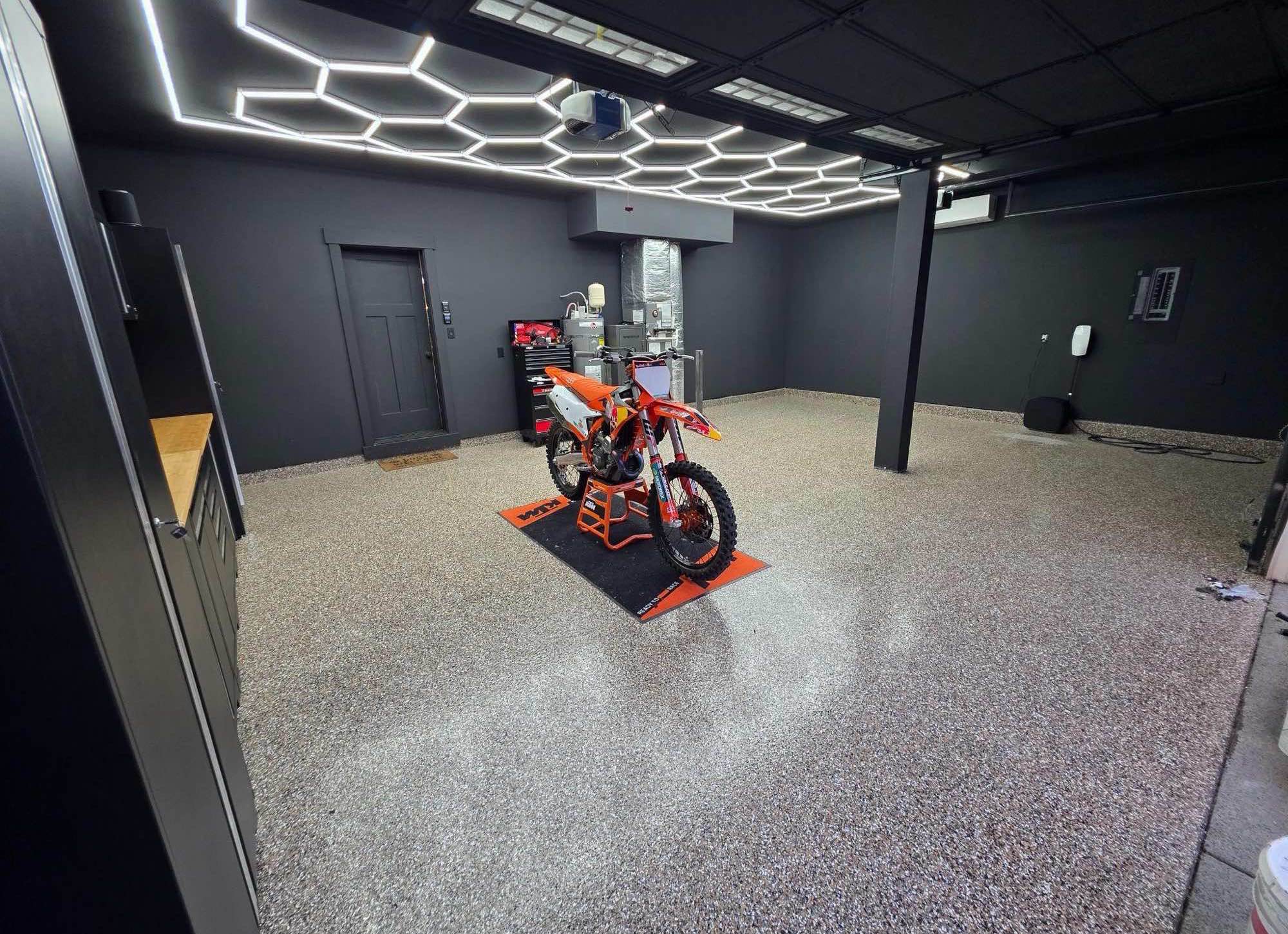 The same garage finished — charcoal walls, a tan flake floor and a hexagon LED ceiling, with a dirt bike on a stand